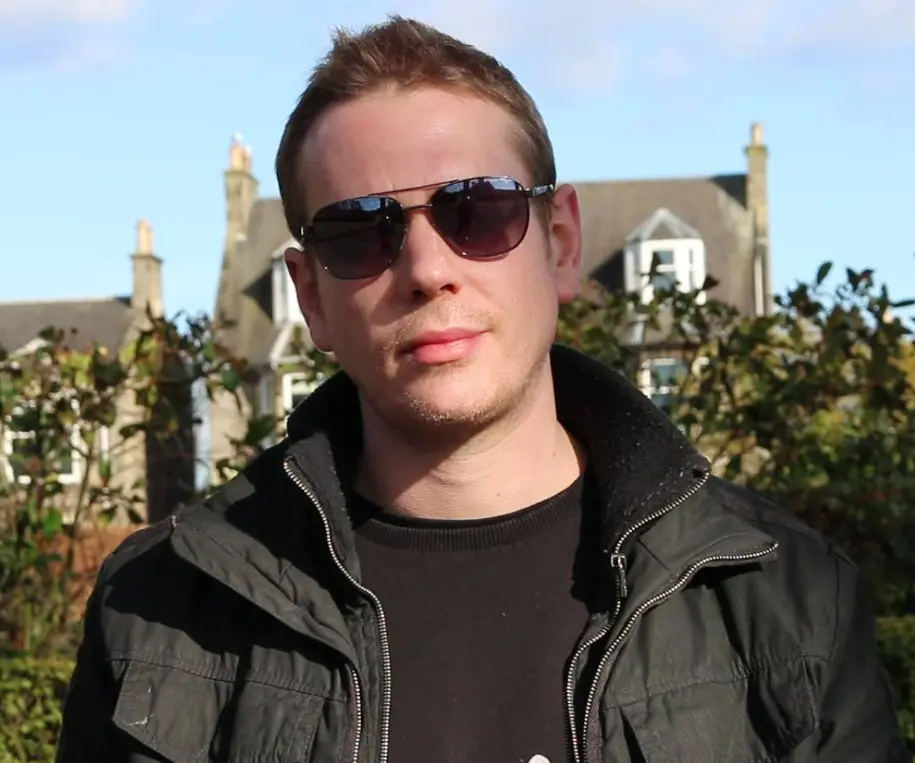 A person in a black jacket stands outdoors, with a backdrop of green foliage and a house under a clear blue sky.